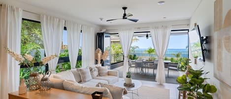 Un salon moderne comprenant un canapé, une table basse et un coin repas, avec de grandes fenêtres offrant une vue sur l’extérieur.