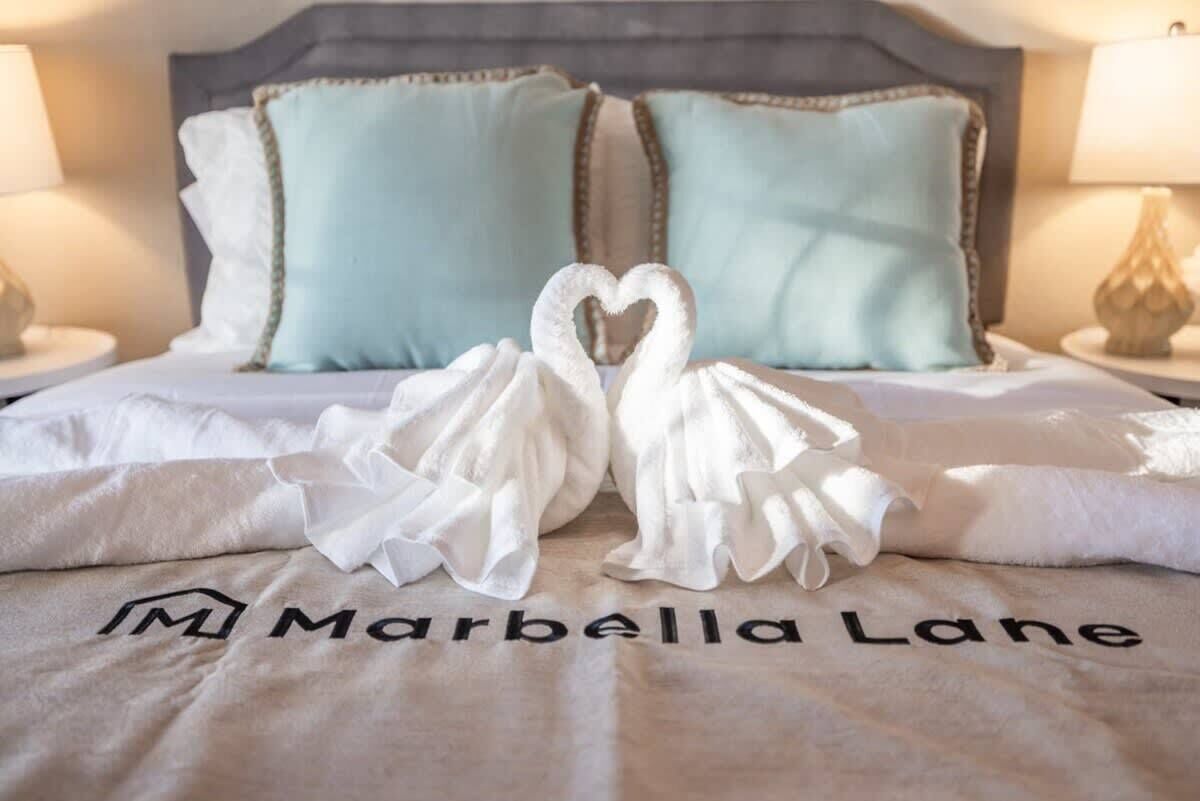 A bed with a white bedspread featuring a heart-shaped towel arrangement and the text "Marbella Lane".