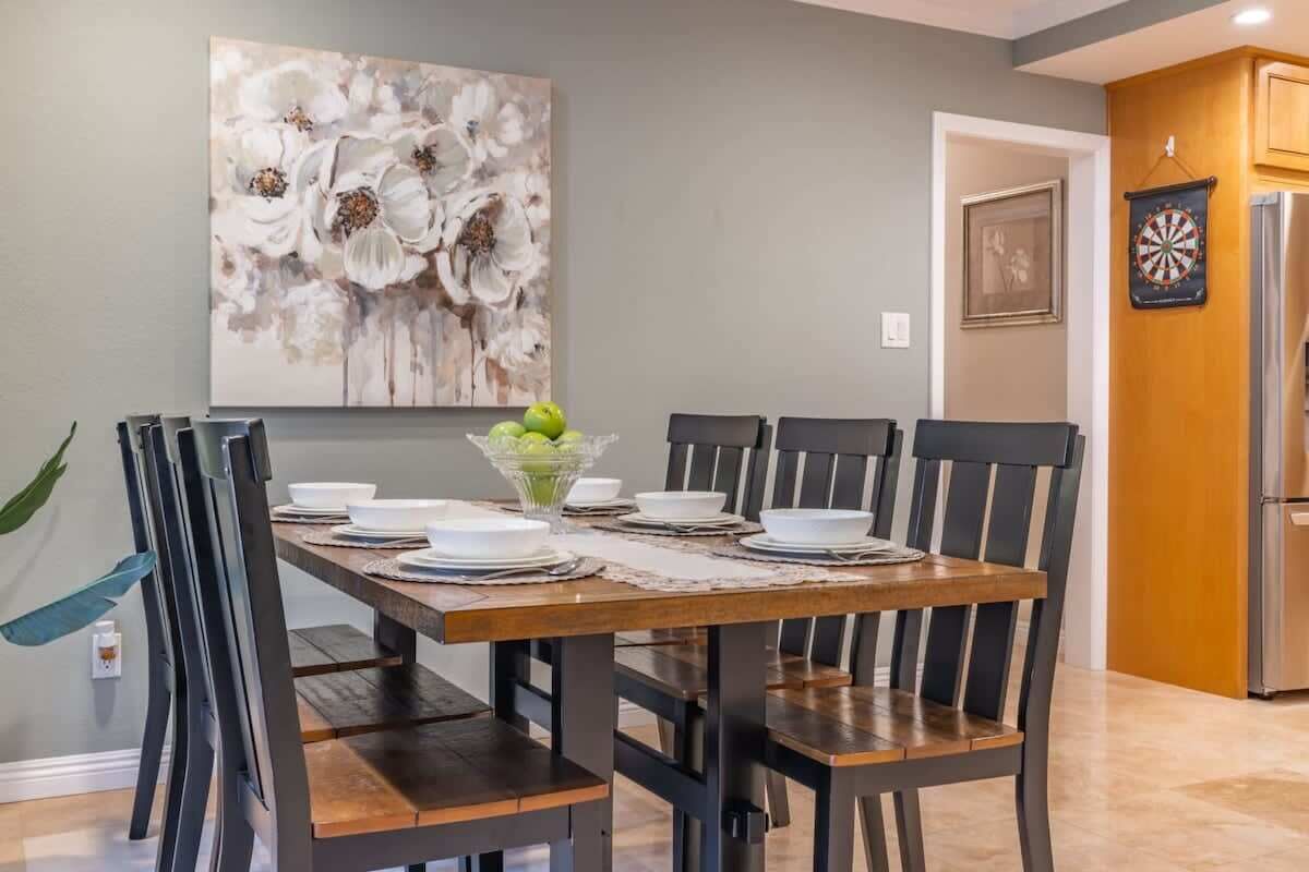 A dining area with a wooden table set for six, surrounded by chairs. A floral painting is on the wall above the table.