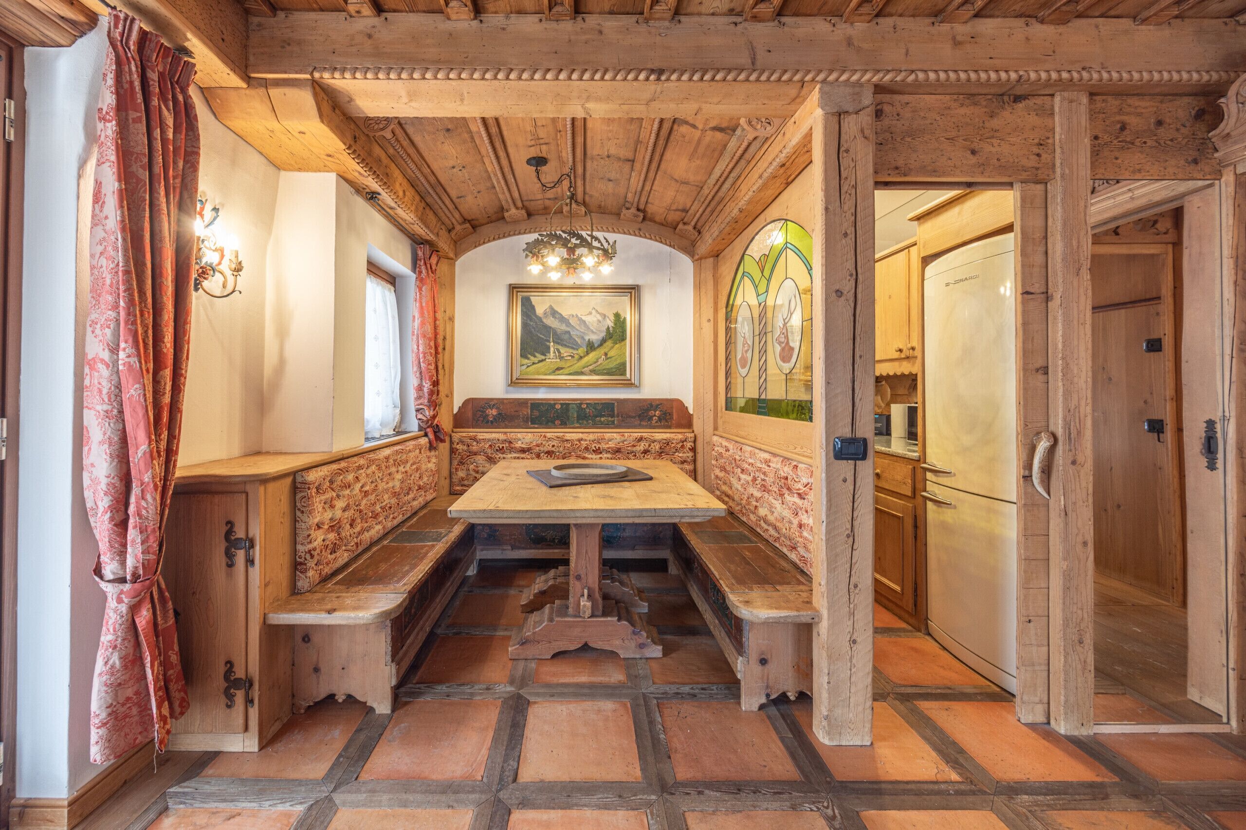 Un interno in legno dallo stile tradizionale, con una zona salotto con panche, un tavolo e una cucina visibile attraverso una porta aperta.