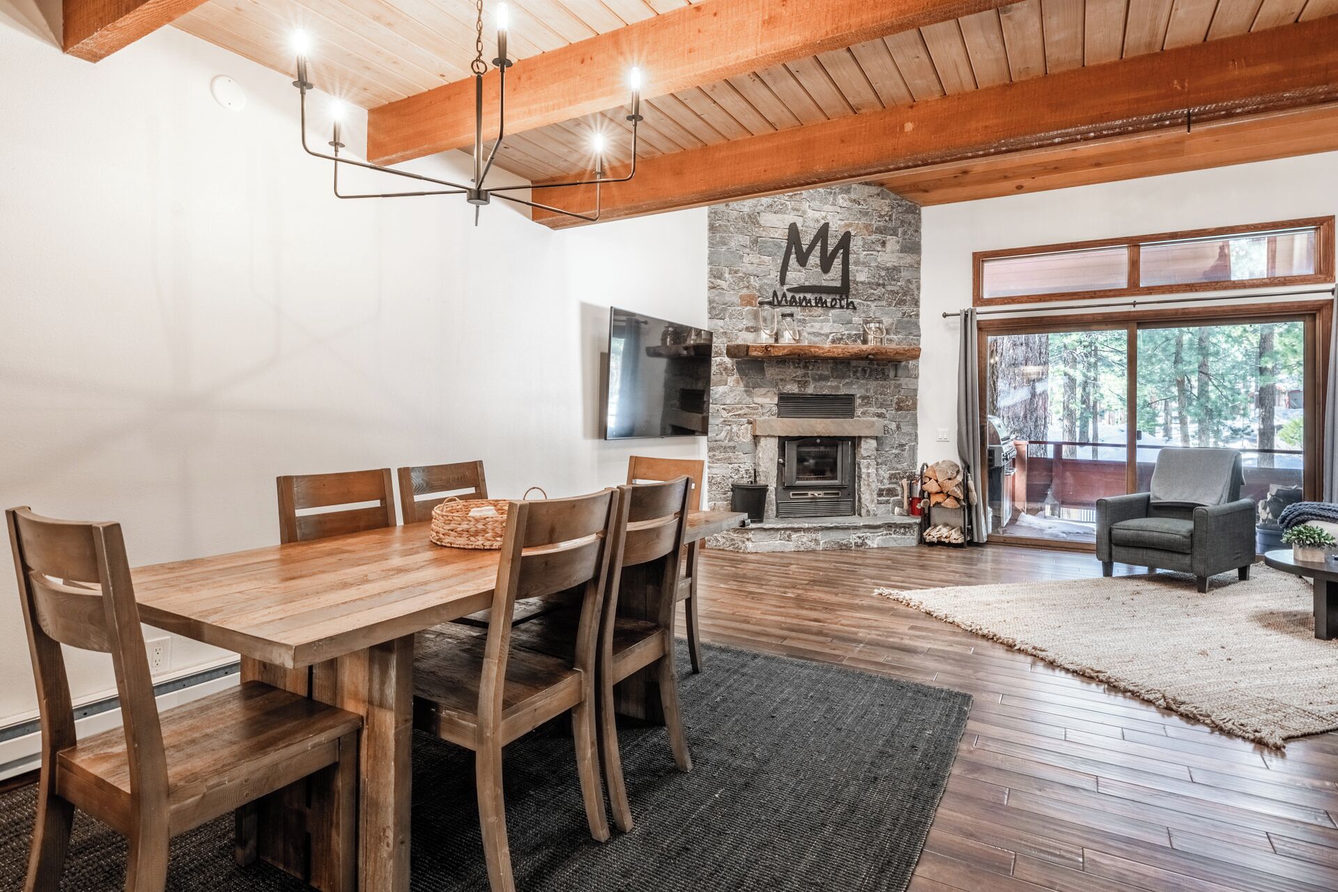 Un vaste espace de vie comprenant une table à manger en bois, des chaises, une cheminée en pierre ornée d’une enseigne « Mammoth », un téléviseur et une grande fenêtre donnant sur la forêt.