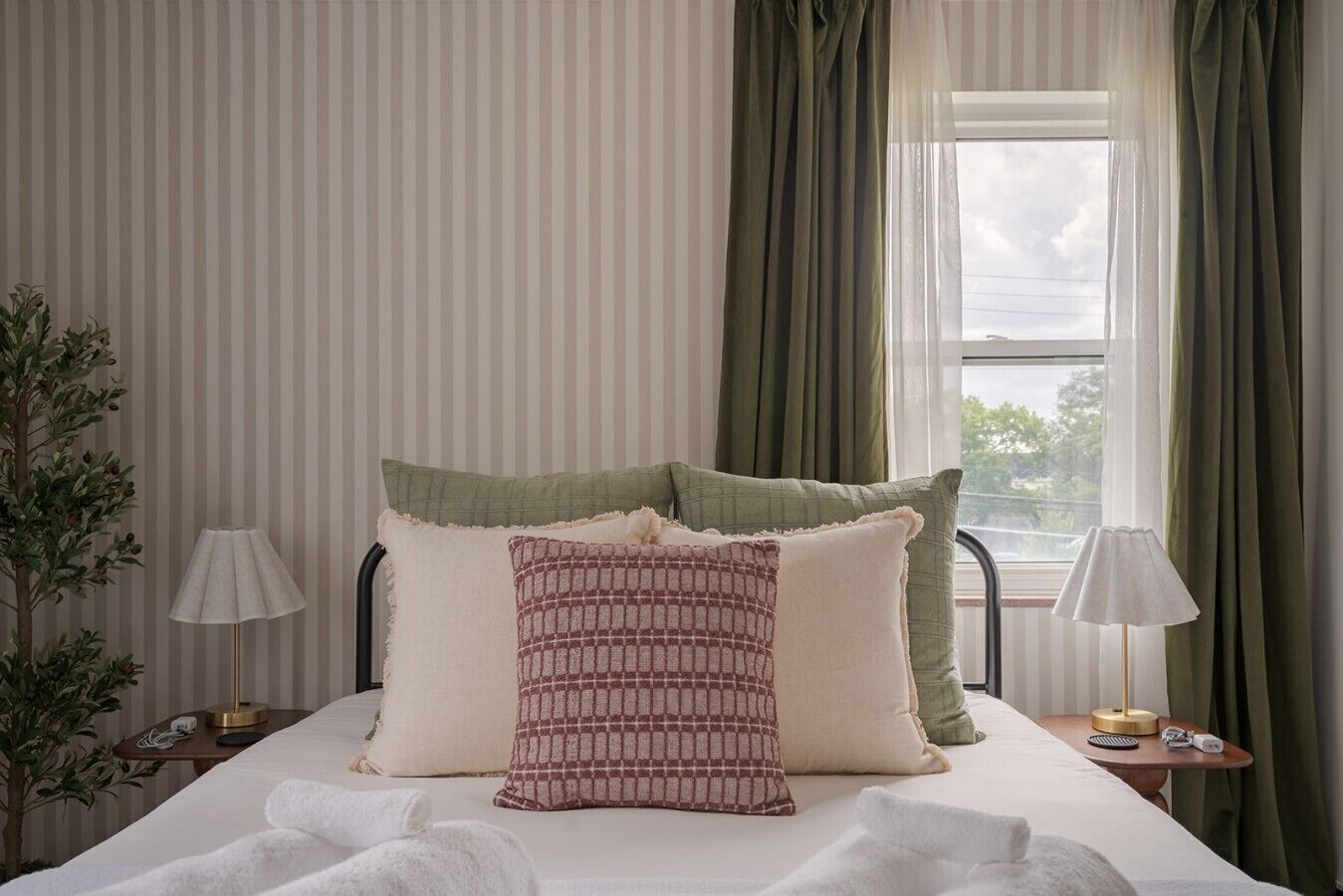 Een bed met kussens, een nachtkastje met een lamp, een plant en een raam met gordijnen.
