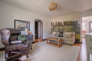 Un salon comprenant un fauteuil en cuir, un canapé, une table basse en bois, un tapis à motifs et un grand tableau représentant un paysage accroché au mur.