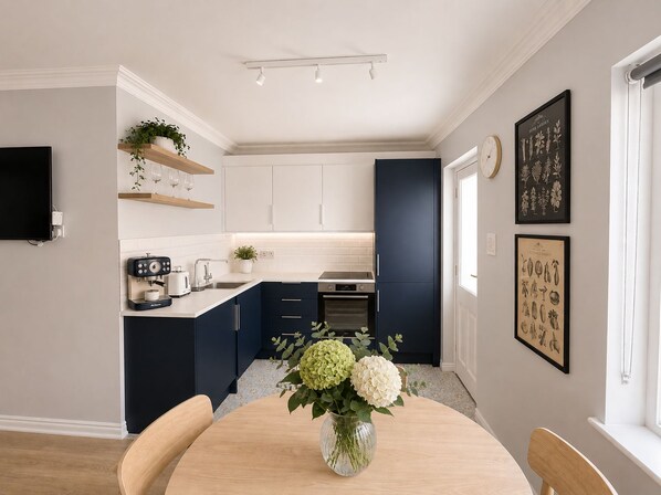 A modern kitchen with white and navy blue cabinets, a coffee machine, and a dining table with a vase of flowers.