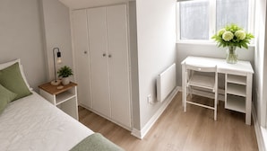 A bedroom with a sloped ceiling, a bed with a green blanket, a bedside table with a lamp and plant, a white desk with a vase of flowers, and a window with curtains. - Super City Centre Location - Sleeps 5 (Galway)