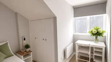 A bedroom with a sloped ceiling, a bed with a green blanket, a bedside table with a lamp and plant, a white desk with a vase of flowers, and a window with curtains.
