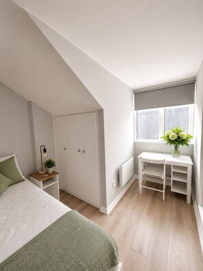A bedroom with a sloped ceiling, a bed with a green blanket, a bedside table with a lamp and plant, a white desk with a vase of flowers, and a window with curtains.
