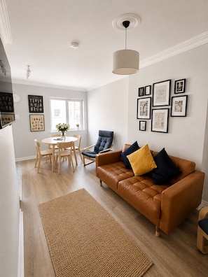 A modern living room with a brown leather sofa, a dining table with chairs, and a large window.