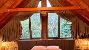 Une chambre avec un grand lit, des murs en pierre et une vue sur les arbres, visible à travers deux fenêtres.