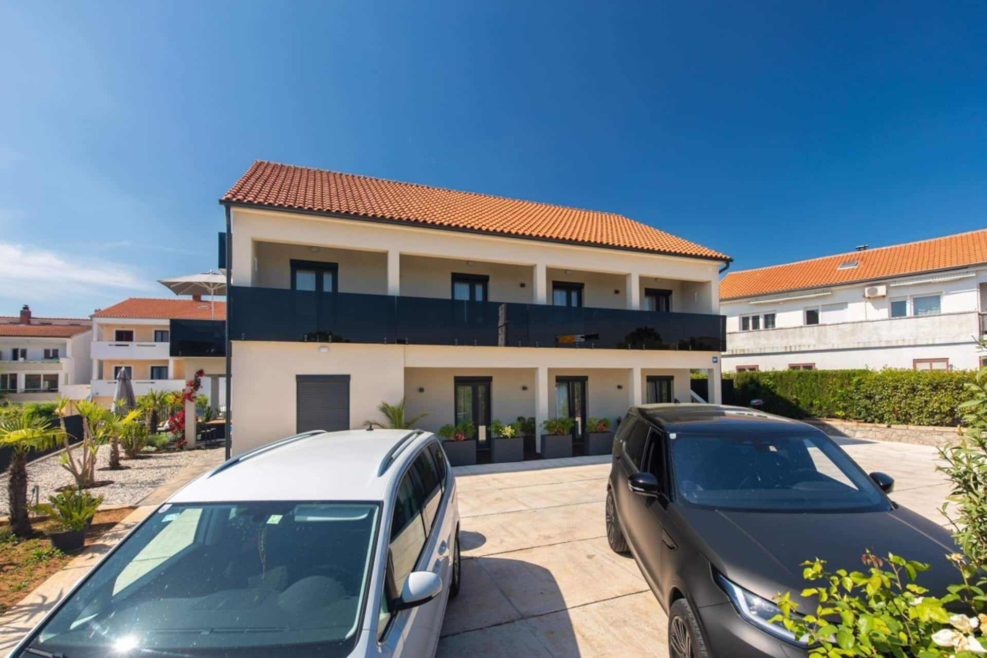 A two-story residential building with a tiled roof, a balcony on the upper floor, and a parking area with two cars.