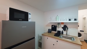 Een compacte keuken met een koelkast, magnetron en koffieapparaat. Er is een spoelbak, een klein aanrecht en een wasmachine op de achtergrond.