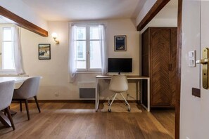 Ein Zimmer mit Schreibtisch, Stuhl und Fernseher. Zwei Fenster mit Vorhängen, eine Tür mit Holzverkleidung und ein Poster an der Wand.