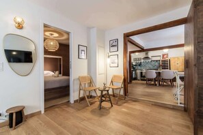 Ein modernes Interieur mit Holzboden, einer Essecke mit Stühlen, einem an der Wand montierten Fernseher und einer durch einen offenen Durchgang sichtbaren Küche.