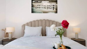 Una cama bien hecha con ropa blanca, un jarrón con flores, una bandeja con croissants y una taza, y una obra de arte que representa una escena de playa.