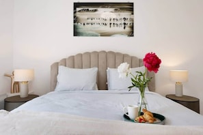 Un lit bien fait, avec du linge de lit blanc, un vase rempli de fleurs, un plateau avec des croissants et une tasse, et une œuvre d’art murale représentant une scène de plage.