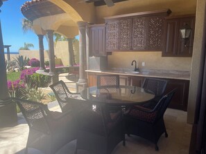 Un área de cocina al aire libre con una mesa redonda de vidrio y sillas de mimbre, rodeada de plantas decorativas y una pérgola.