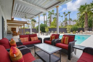 Un patio techado al aire libre con mobiliario de mimbre, una piscina y palmeras.