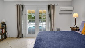 A bedroom with a bed, a bookshelf, an air conditioning unit, and a view of an outdoor swimming pool through a glass door. - Casa Chiquita: HUGE Backyard, Heated Pool & Spa (Palm Springs)