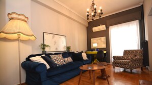 A living room with a blue sofa, a floor lamp, a patterned armchair, a small round table, and a chandelier. - Bibelot Acropolis (Athens)