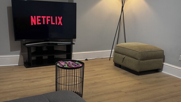 Un saló amb una televisió de pantalla plana que mostra 'NETFLIX', un sofà gris, una taula rodona petita amb un cistell, una làmpada de peu i un otomanet beix.