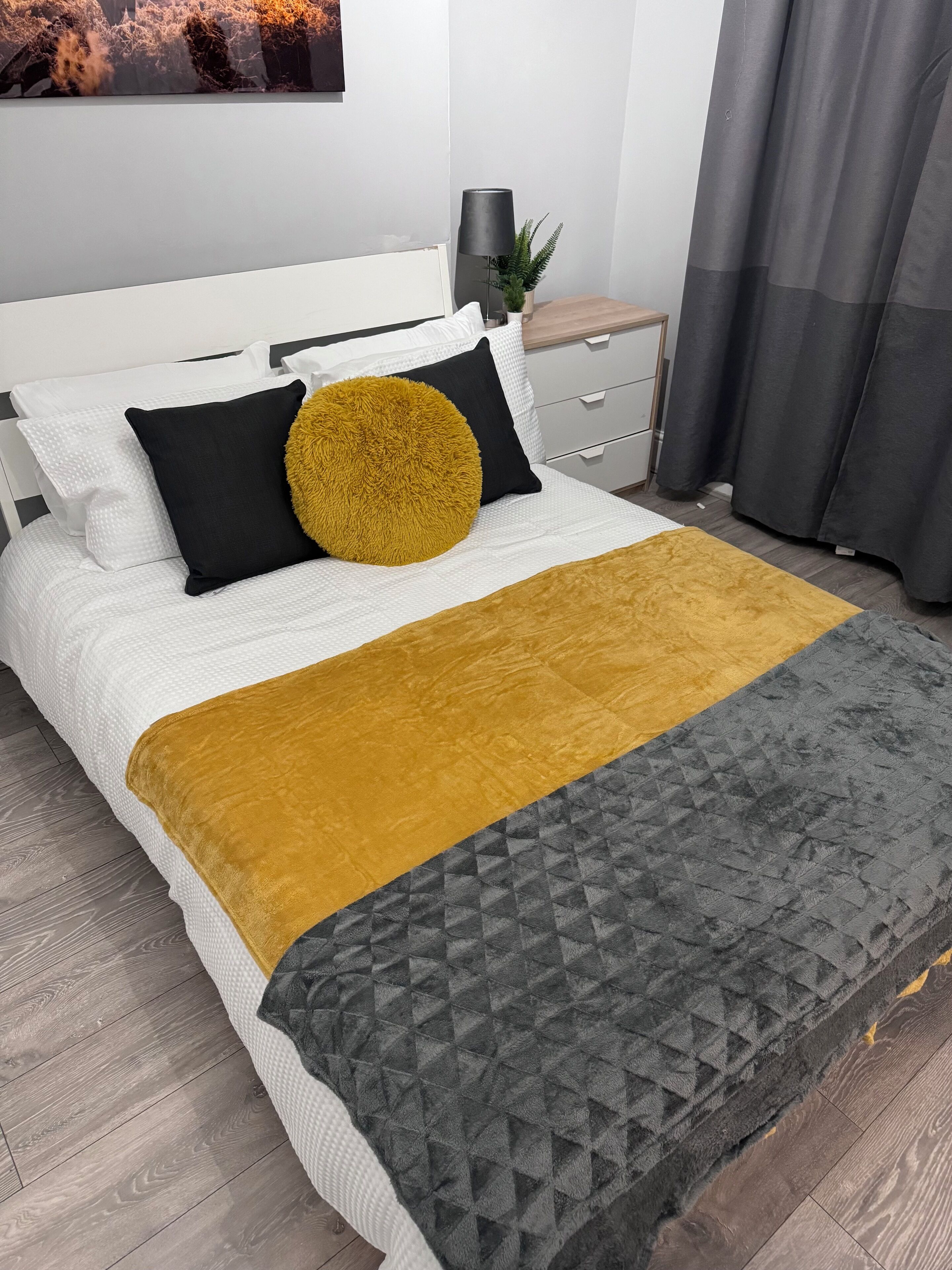 A neatly made bed with a white headboard, a mustard-colored throw blanket, and a grey quilted blanket.