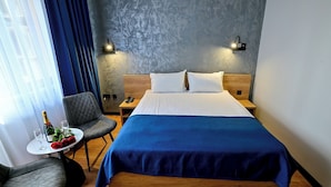 A hotel room with a bed, bedside tables, a chair, a small table with a bottle and glasses, and a view of the outside through a window with curtains.