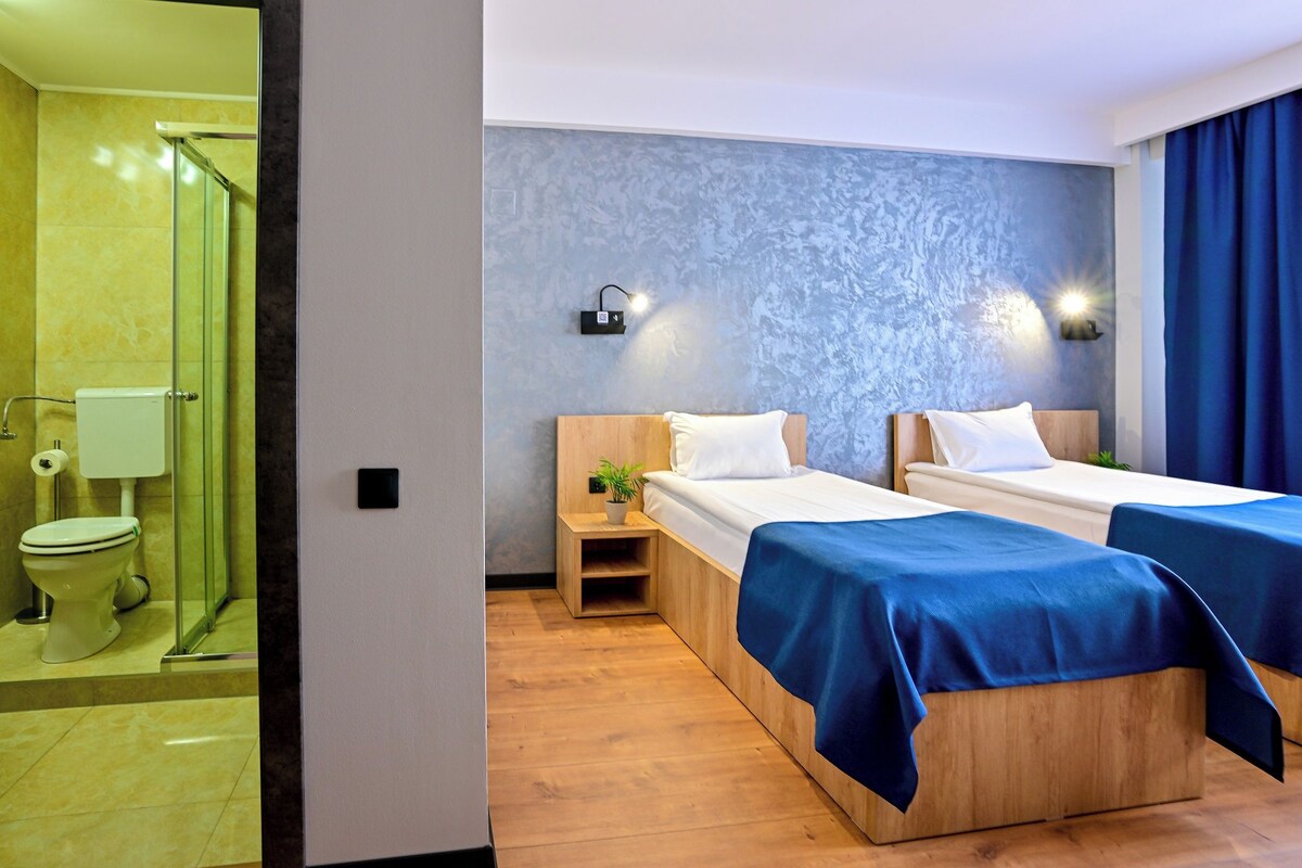 A hotel room with two beds, a shower, and a small bedside table with a lamp.