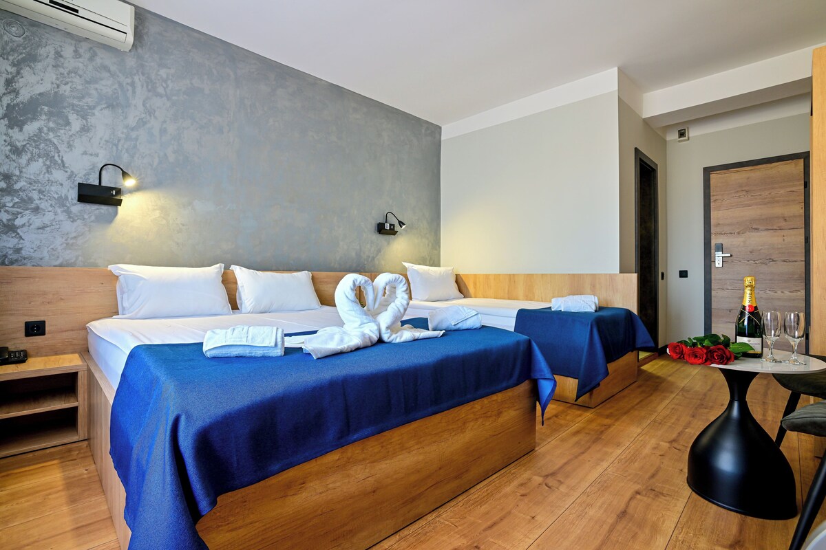 Habitación de hotel con dos camas, piso de madera, lámpara fijada a la pared, puerta con panel de madera y una mesa con una botella y vasos.