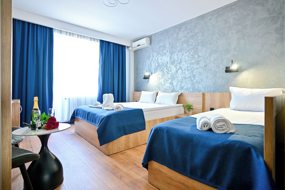 A hotel room with two beds, a small table with a bottle and glasses, and a view of the outdoors.