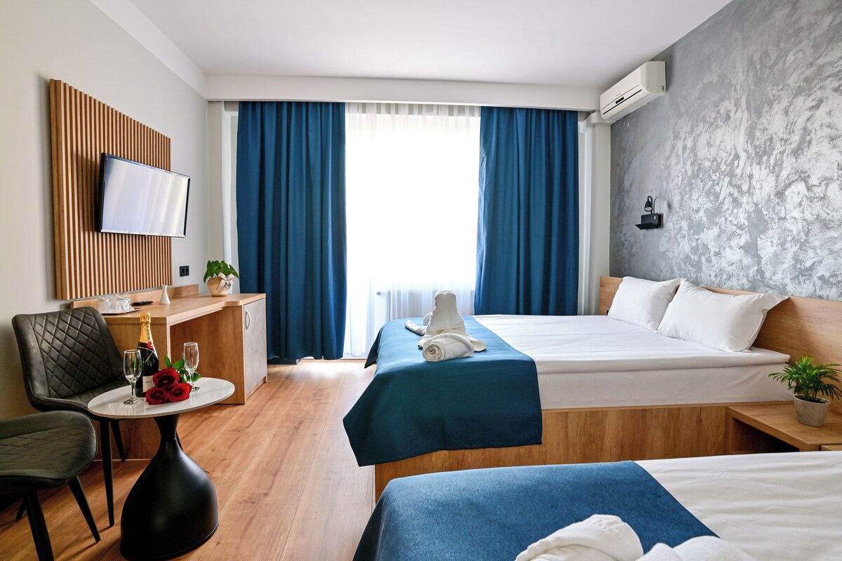 Ein Hotelzimmer mit zwei Betten, einem Schreibtisch mit Stuhl, einem kleinen Tisch mit Flasche und Gläsern sowie einem an einer Holzplatte montierten Fernseher.