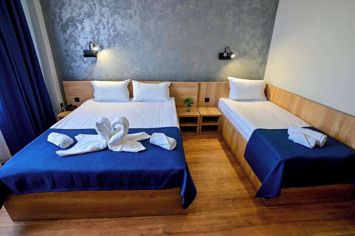 A hotel room with two beds, each with a blue bedspread and white pillows. There are two wall-mounted lights and a small table with a plant.