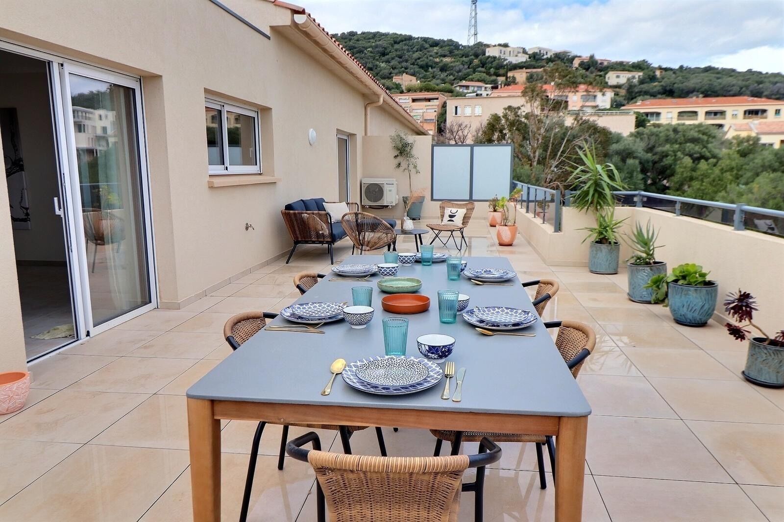 An outdoor dining area with a table set for six, surrounded by wicker chairs, on a terrace with potted plants and a view of the city.