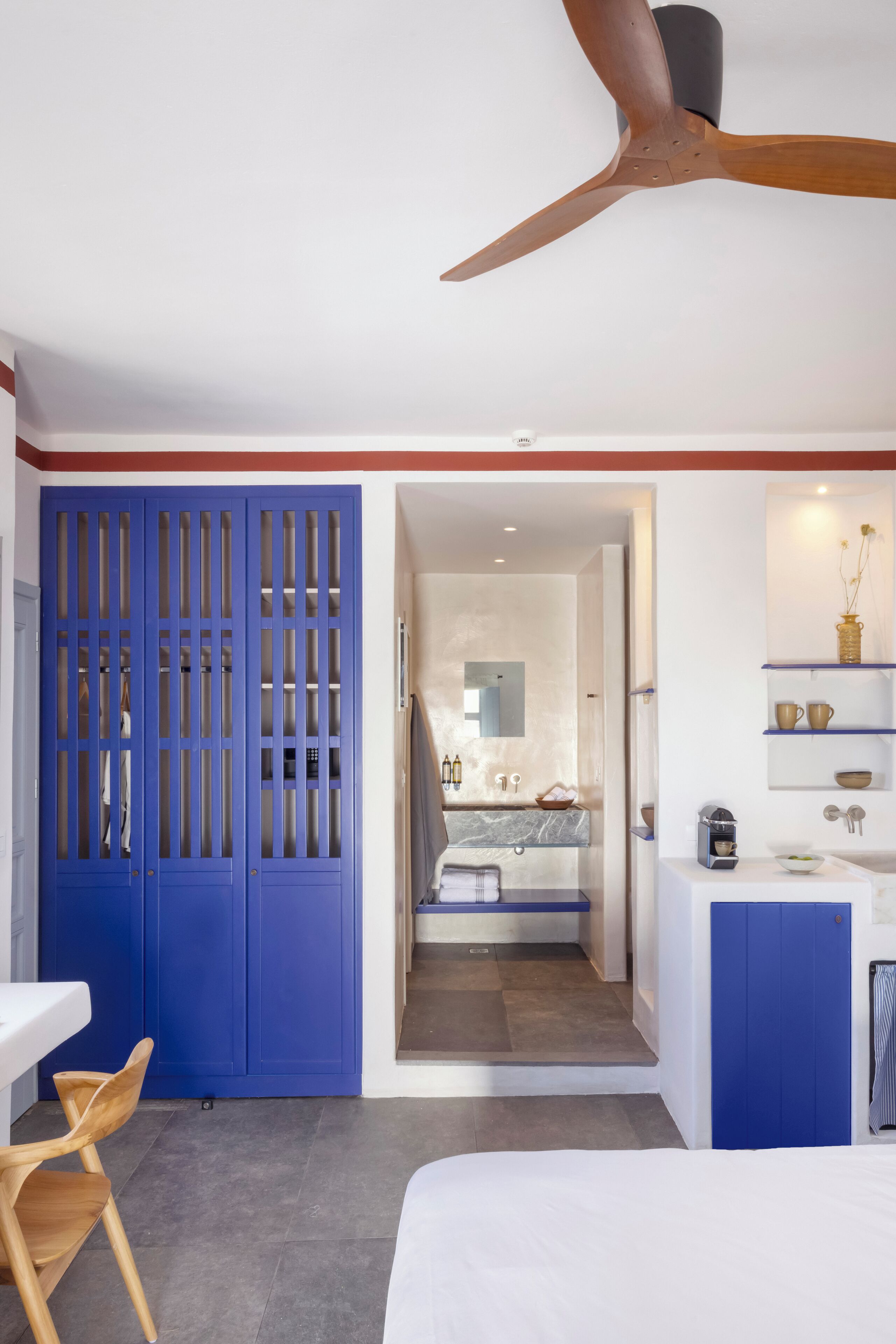 Ein modernes Schlafzimmer mit Bett, Deckenventilator, Badezimmer mit Spiegel und Waschbecken sowie einem blauen Kleiderschrank.