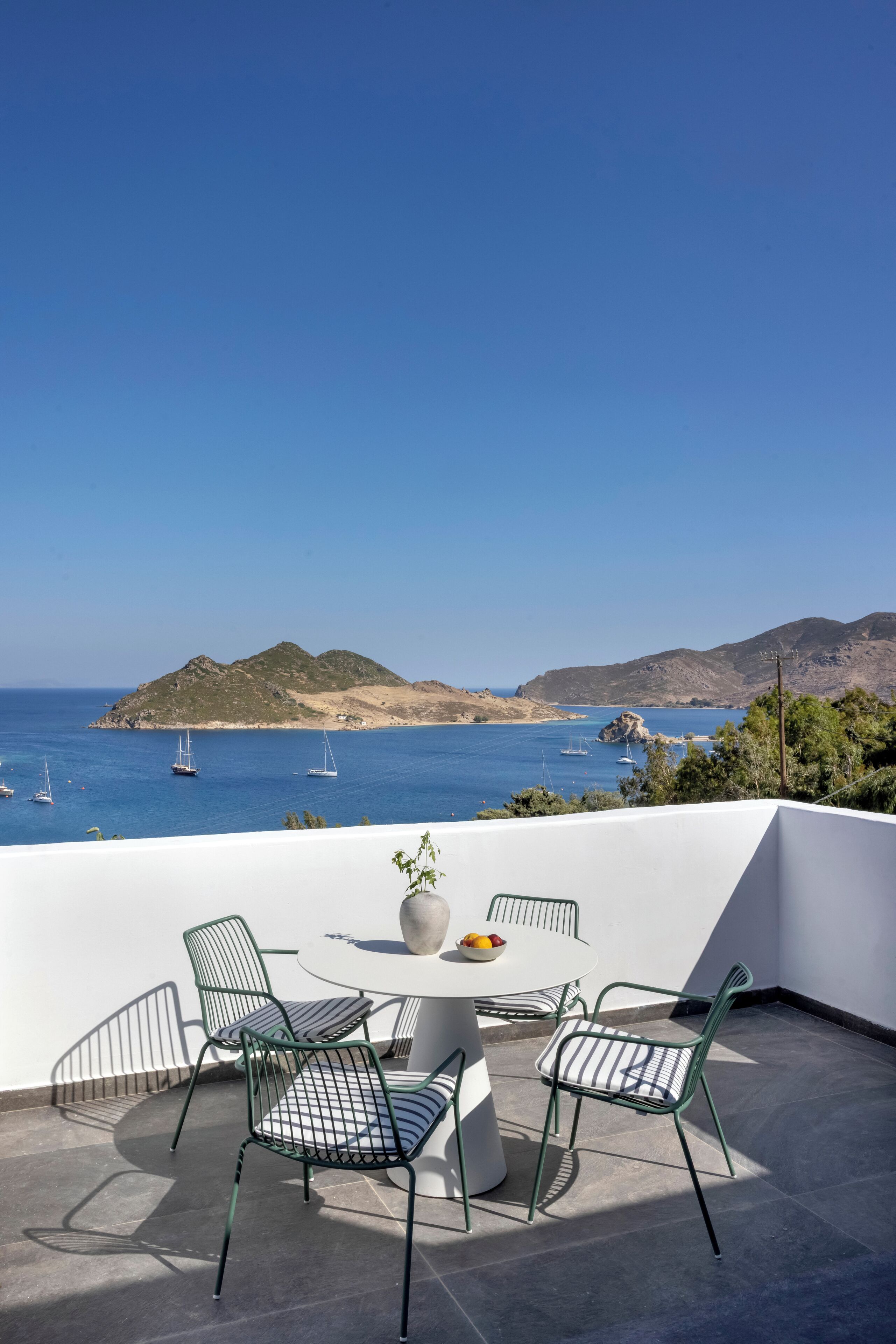 Una terraza con una mesa redonda y cuatro sillas, con vistas a un paisaje costero con barcos y colinas.