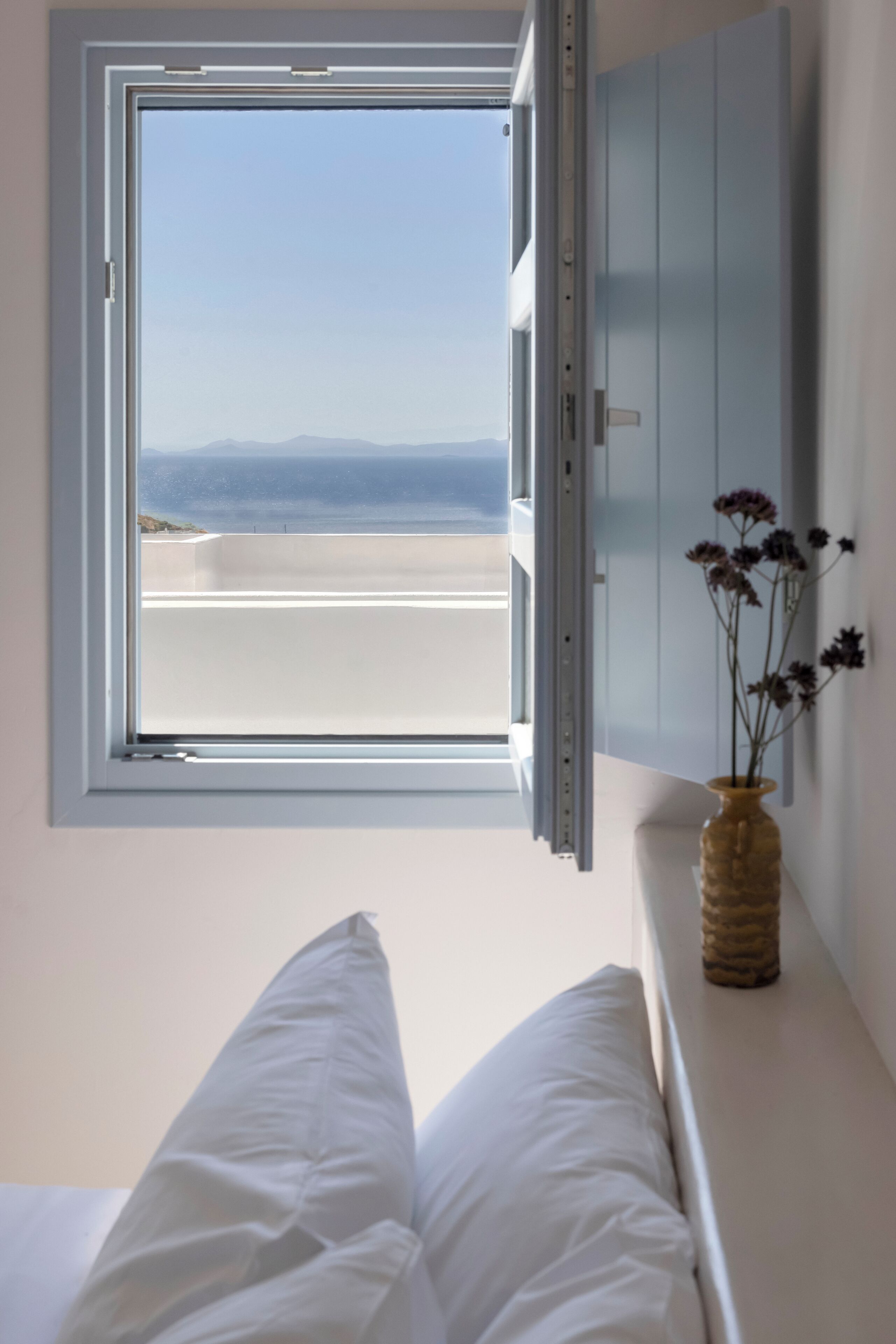A bedroom with a large window, a bed with white bedding, and a vase with flowers on a bedside table.
