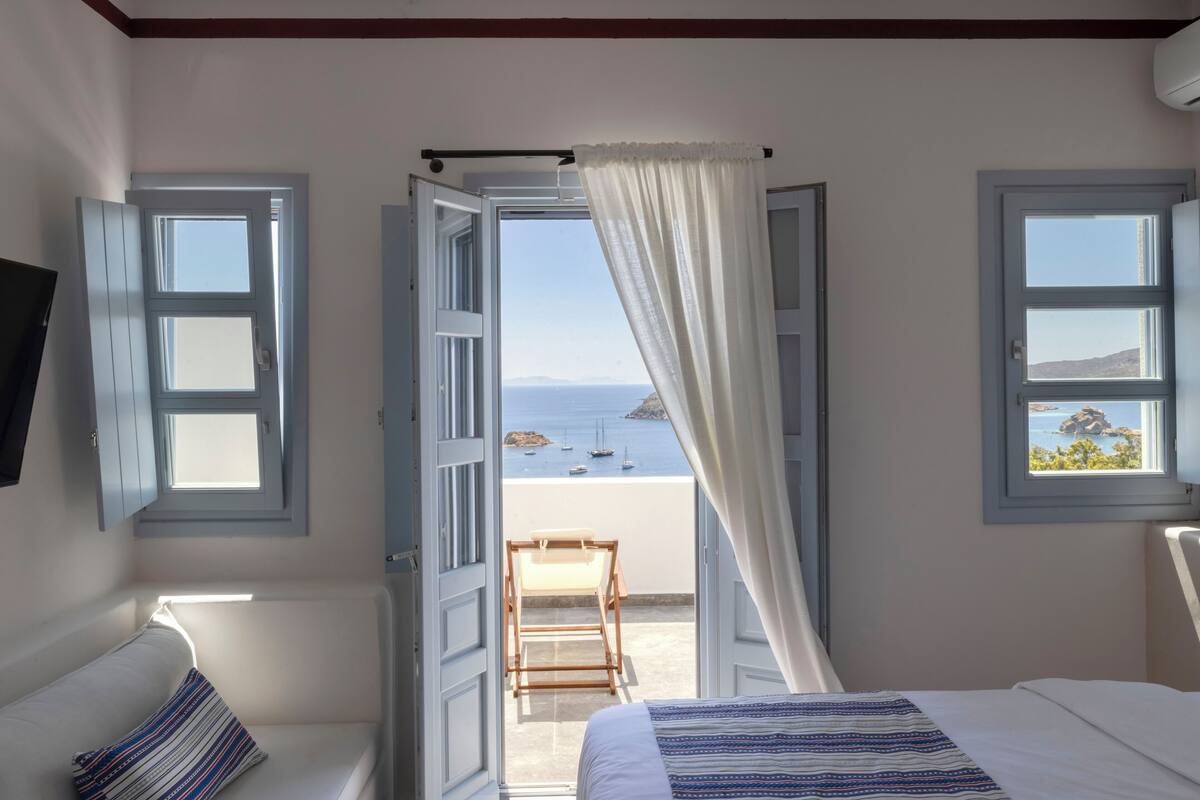 Een slaapkamer met een bed, een stoel en uitzicht op zee door open ramen.