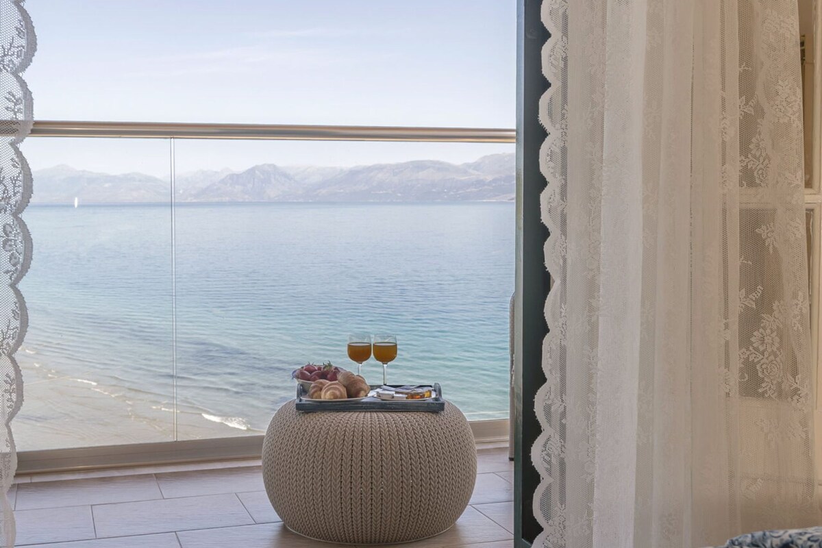 A bedroom with a view of a lake, a bed, a bedside table with glasses and pastries, and a woven ottoman.