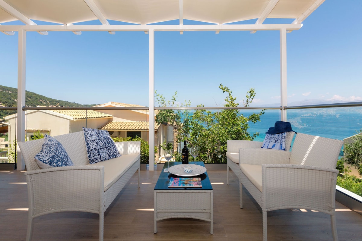 A covered outdoor seating area with wicker furniture, a table with a magazine, and a view of the sea.