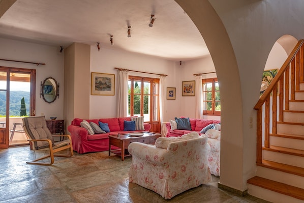 A spacious living room with a red sofa, a floral-patterned sofa, and a wooden rocking chair. There are large windows with curtains, a wooden staircase, and a view of the outdoors. - Alisachni Apartments (Corfu)