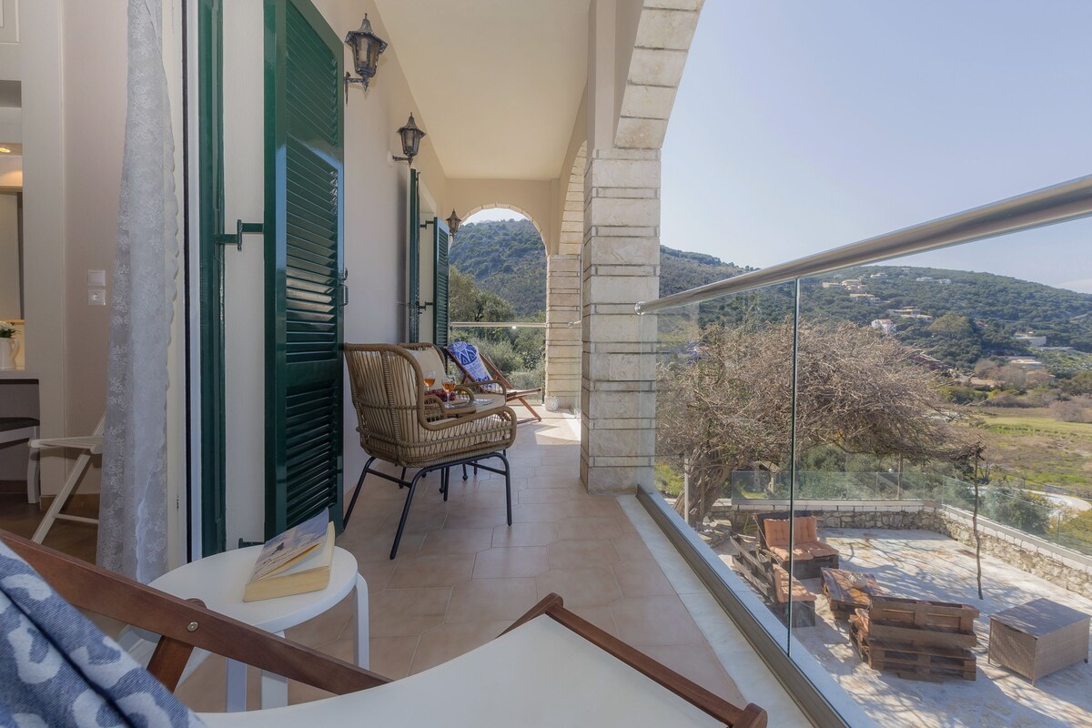 A balcony with a view of a hilly landscape, wicker furniture, and a glass railing.