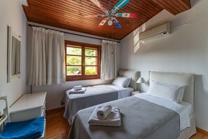 A room with two beds, a ceiling fan, an air conditioning unit, a window with curtains, and a blue towel on a bench. - Alisachni Apartments (Corfu)