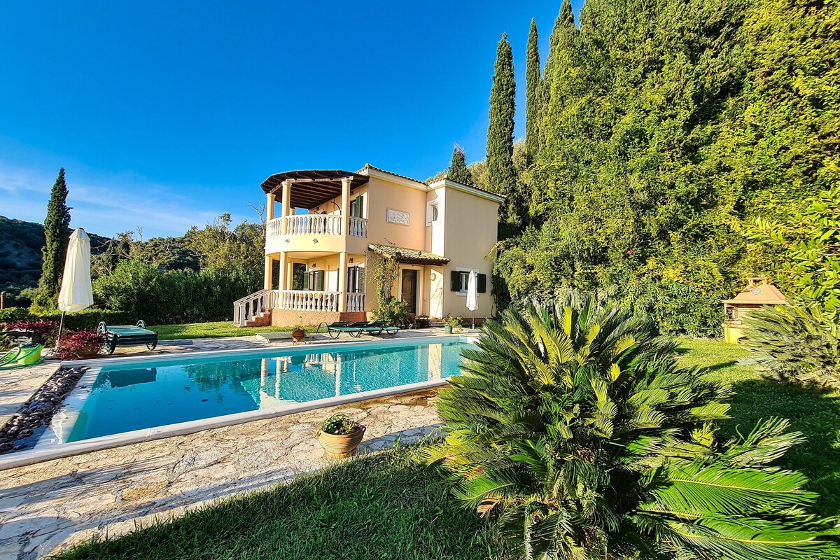 A two-story house with a pool, surrounded by lush greenery and a clear blue sky.