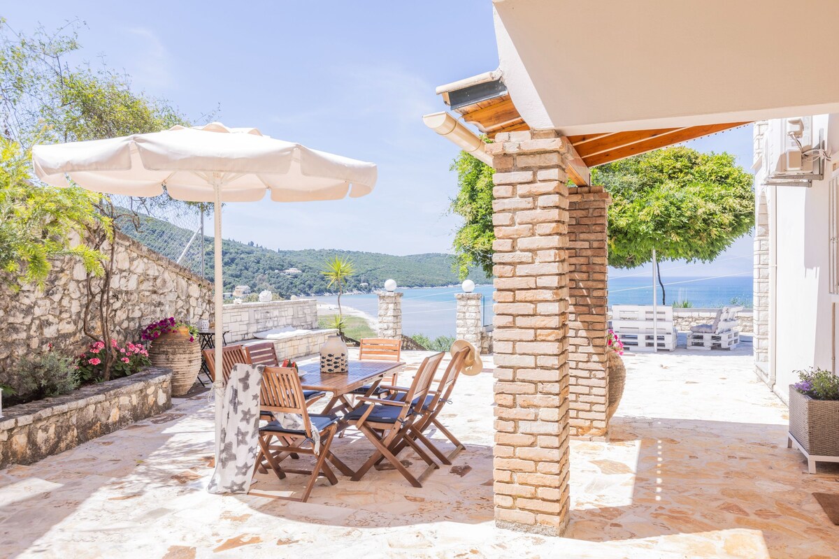 An outdoor patio with a table and chairs, a white umbrella, and a view of the sea.
