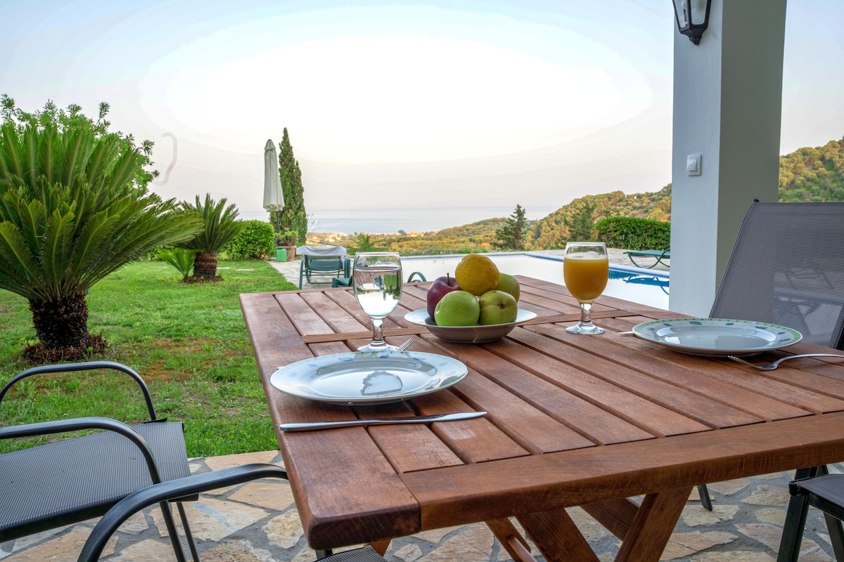 A wooden table with plates, cutlery, a bowl of fruit, and glasses of juice, set against a scenic view of a swimming pool and hills.