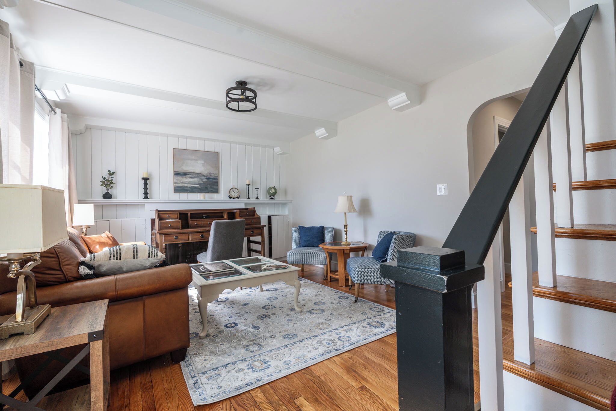 A spacious living room with a sofa, coffee table, and chairs, a kitchen area with white cabinets, and a staircase.