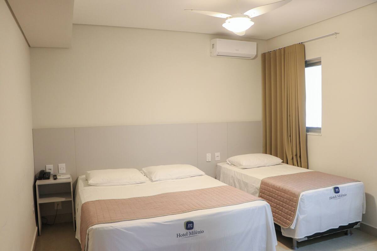 Ein Hotelzimmer mit zwei Betten, einem Nachttisch, einem Deckenventilator, einer Klimaanlage und einem Fenster mit Vorhängen.