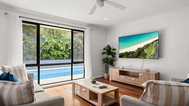 Ein modernes Wohnzimmer mit großem Fenster, einem Flachbildfernseher, Holzmöbeln und Blick auf Pool und Grünflächen.