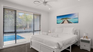 Ein Schlafzimmer mit einem Bett, Nachttischen, einem Deckenventilator, einem Strandbild und Blick auf ein Schwimmbecken durch große Fenster.