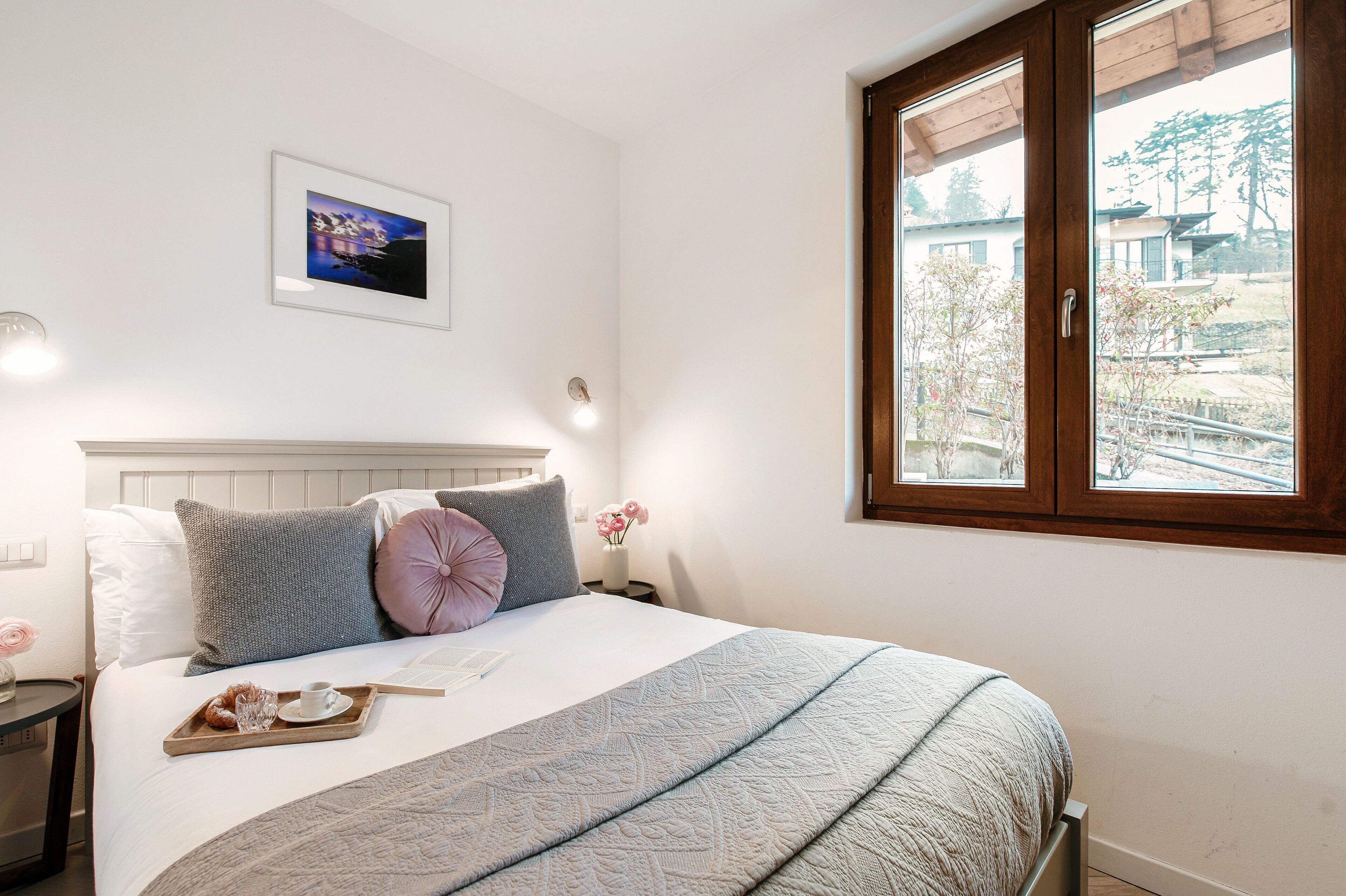 Ein ordentlich bezogenes Bett mit einer Platte Gebäck, ein gerahmtes Bild an der Wand und ein Fenster mit Blick auf Bäume und Gebäude.