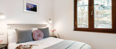 Ein ordentlich bezogenes Bett mit einer Platte Gebäck, ein gerahmtes Bild an der Wand und ein Fenster mit Blick auf Bäume und Gebäude.